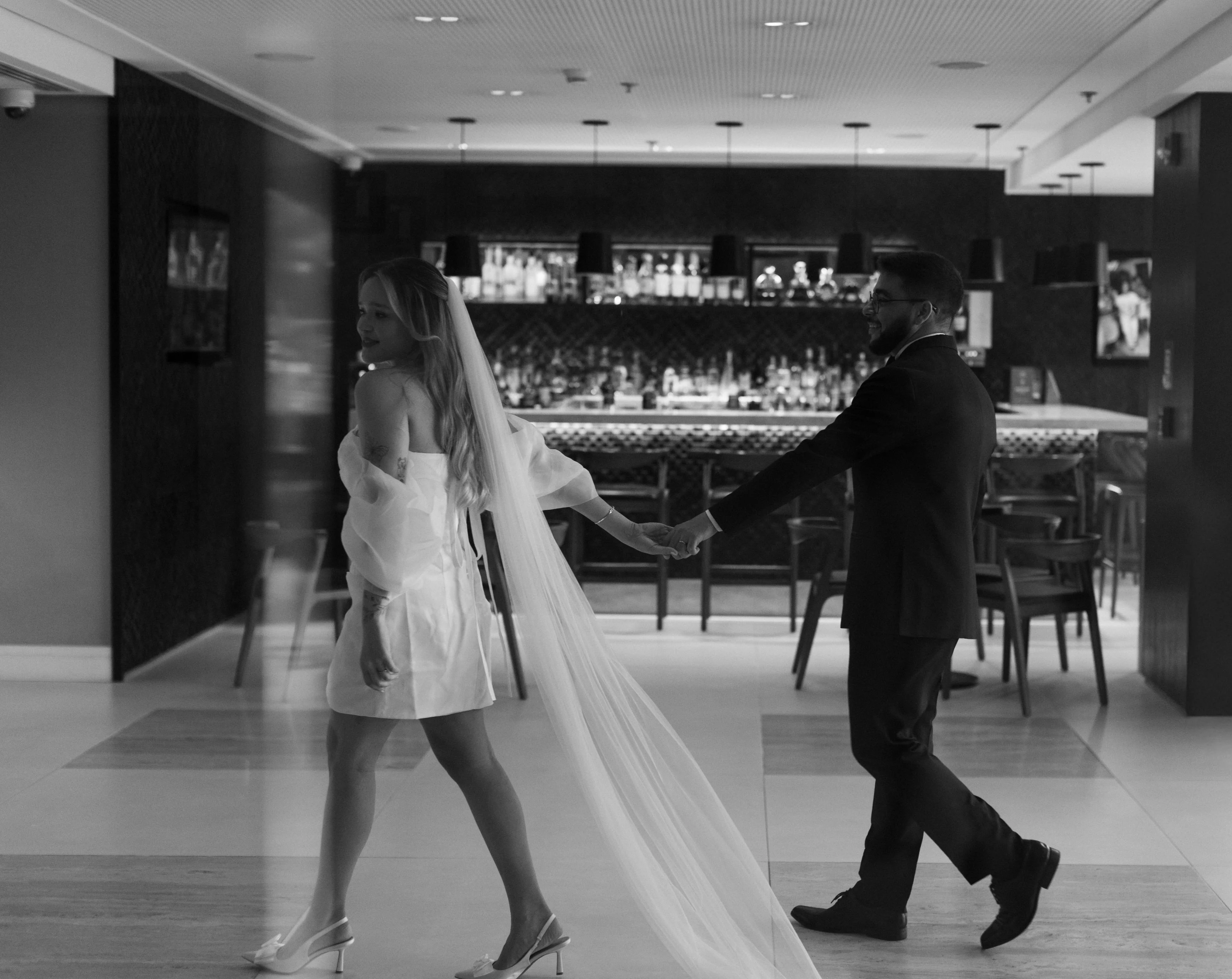 Noiva de vestido curto e véu longo conduz o noivo pelo salão, em preto e branco